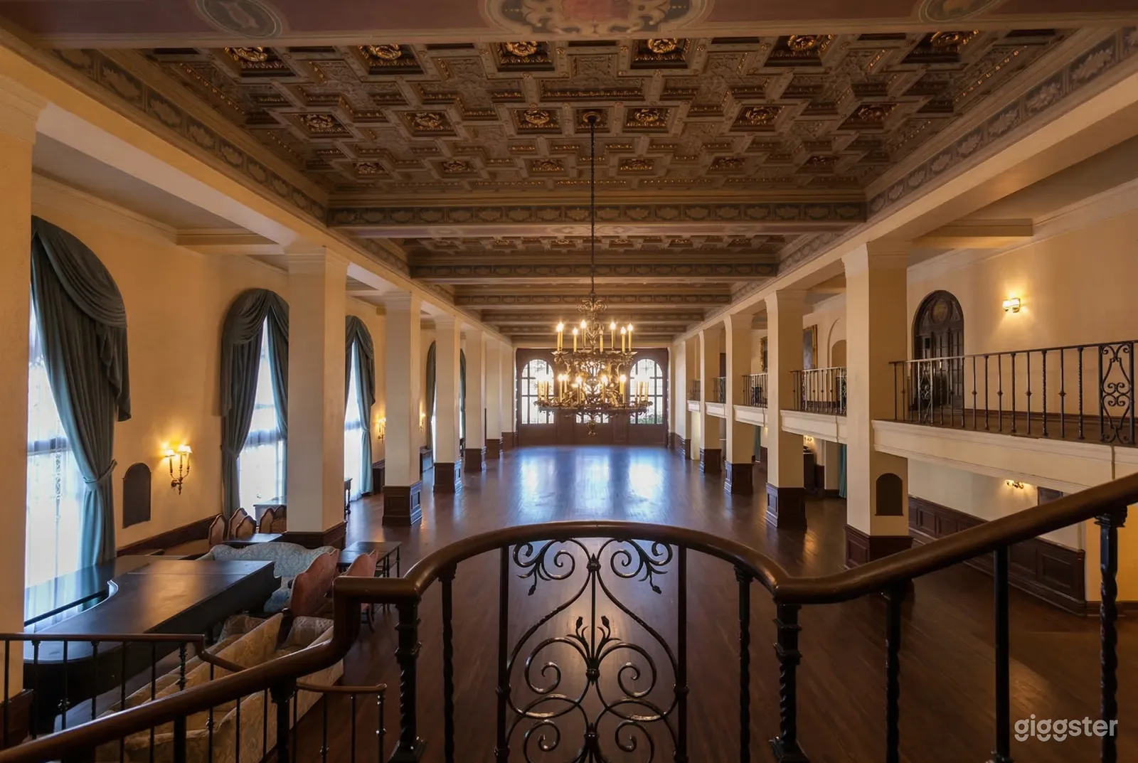 A long room with columns on either side has a shiny brown cement floor, an ornate ceiling, and a single chandelier.