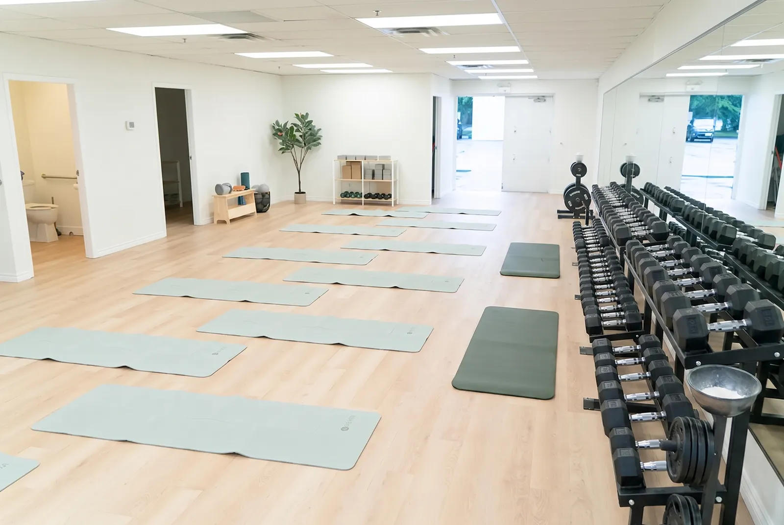 A large and bright training room has workout mats on the floor and rows of hand weights off to the side.