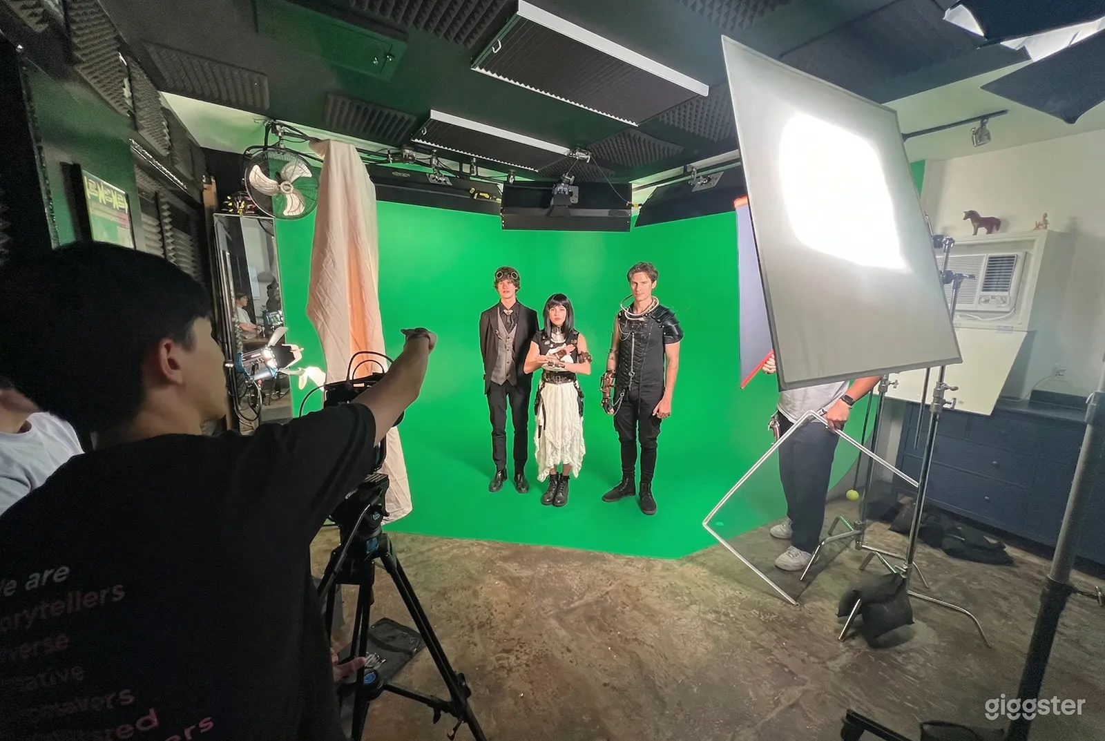 Three people stand in front of a green screen while a director and crew in front of them give instructions.