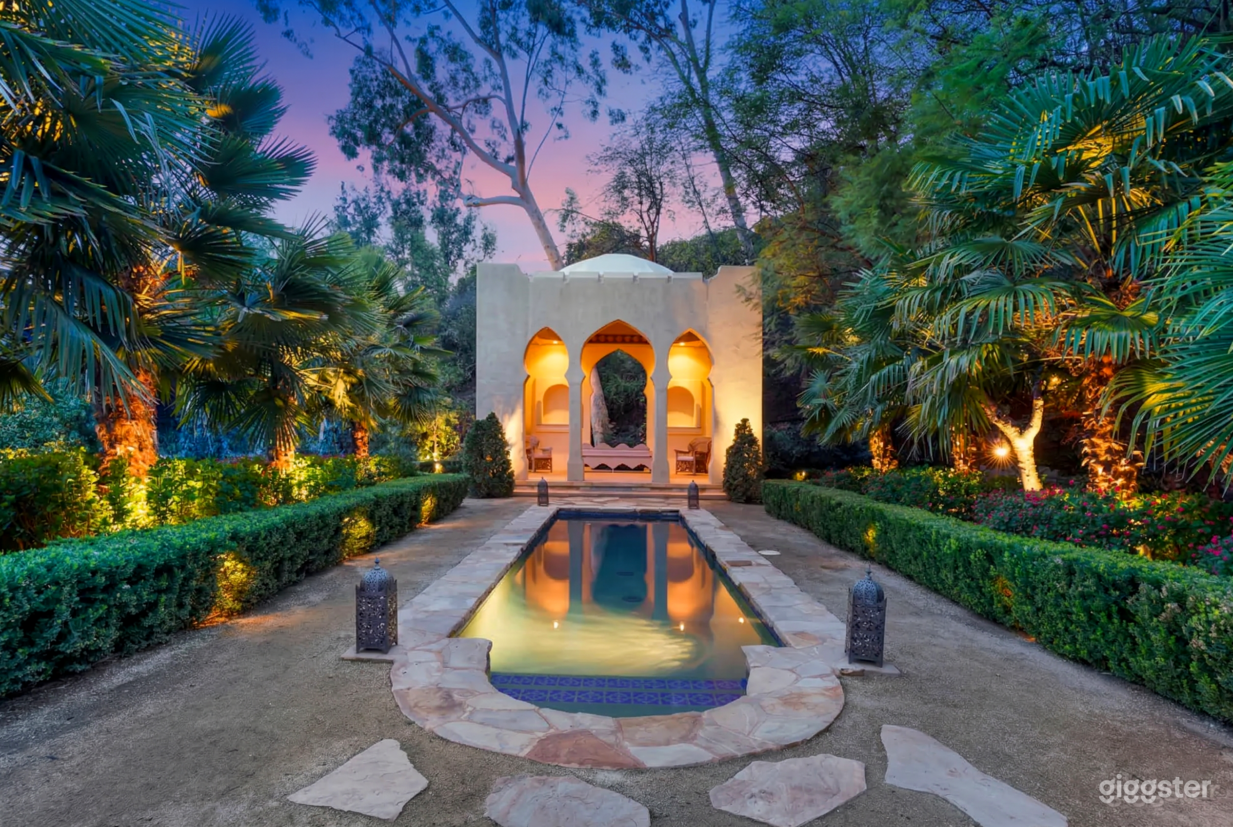 A Mediterranean style cabana is at the head of a long pool surrounded by hedges and tall tropical trees.