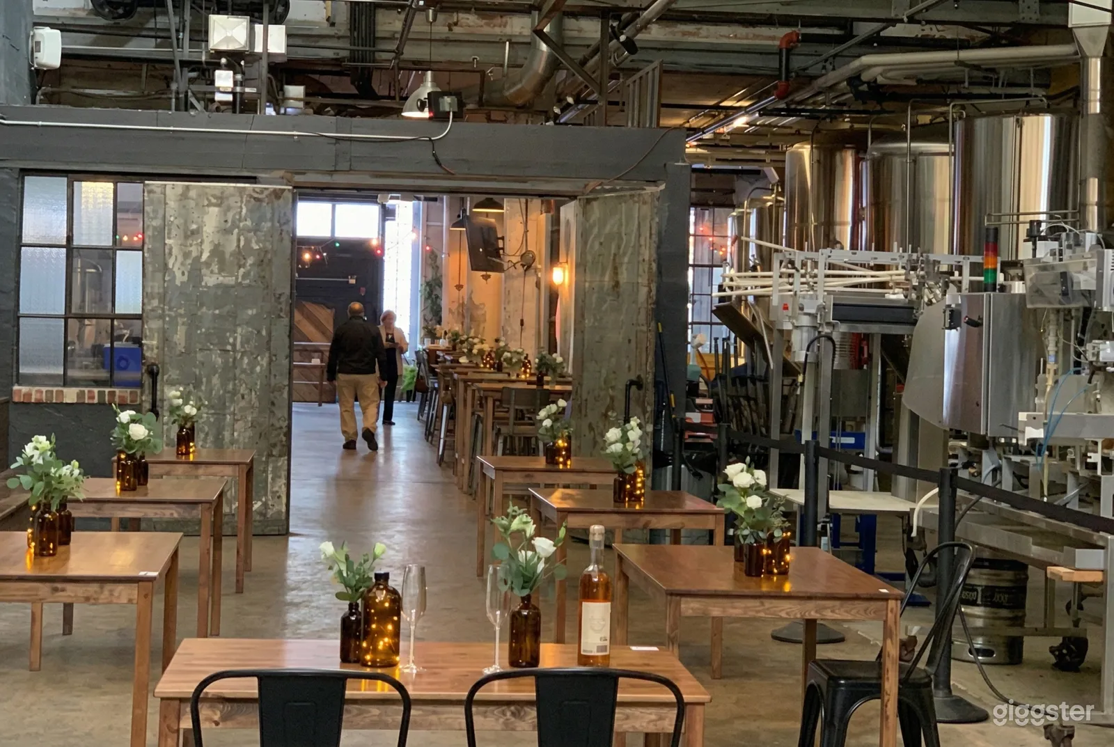 Craft winery steel vessels are to the right of square wooden tables decorated with a floral arrangement and bottle of wine.