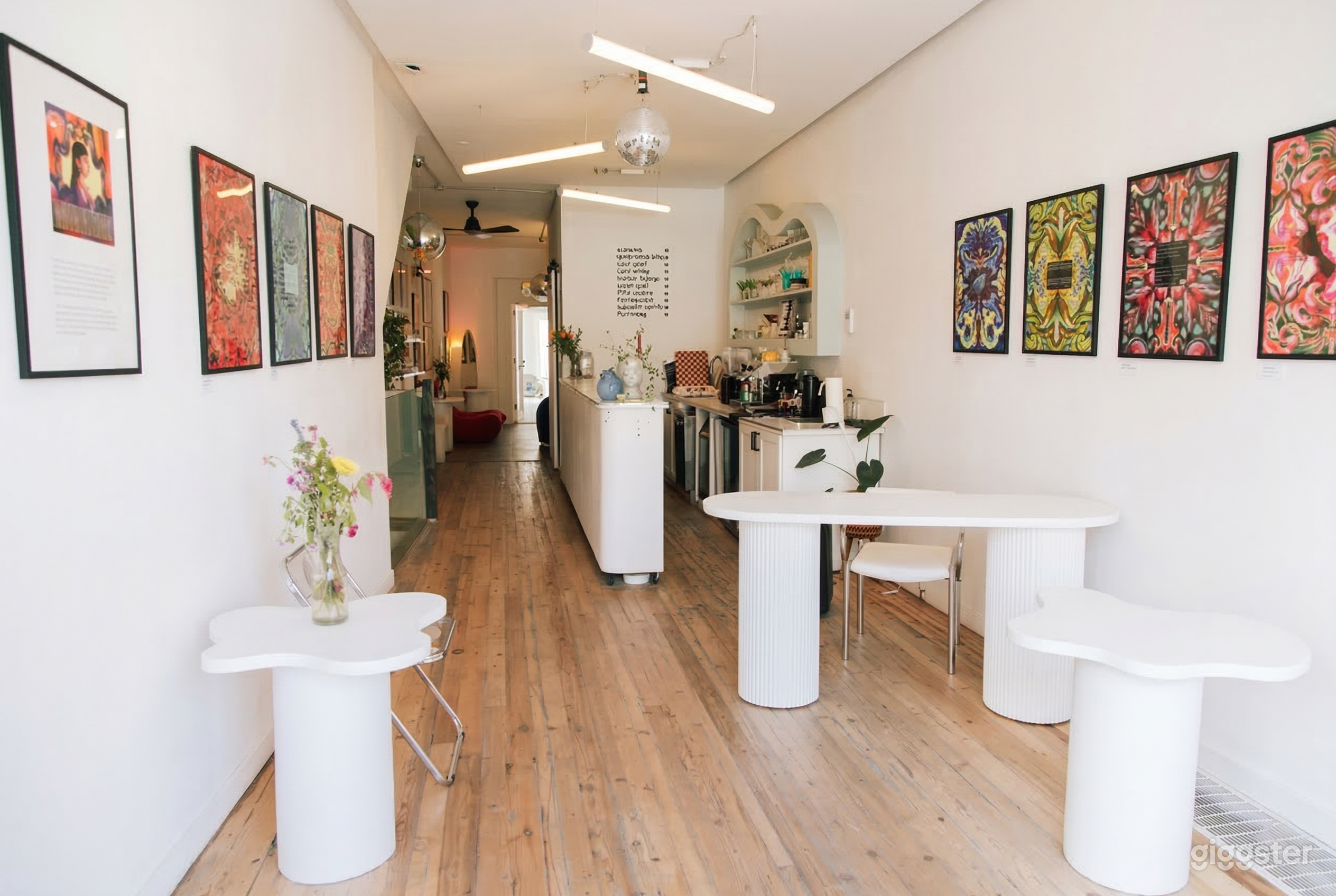 A cafe with white tables and chairs, colorful prints on the wall, and a snack prep area.