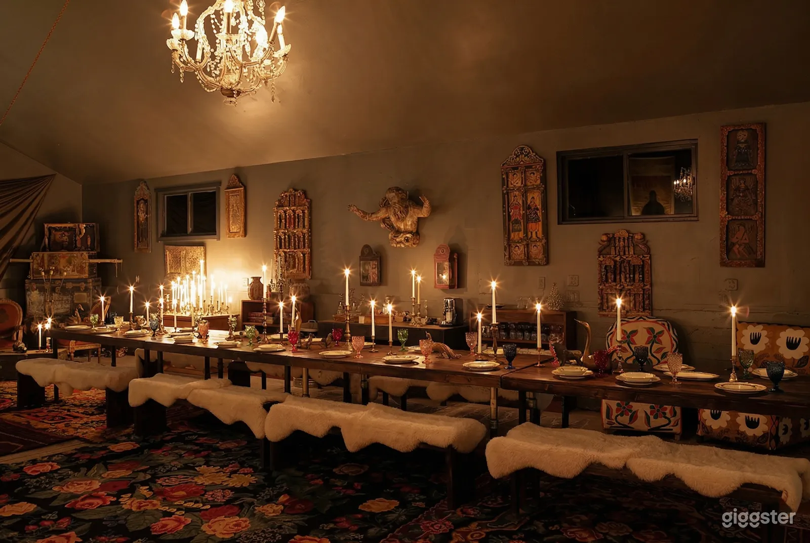 A dim room has walls decorated with wooden art pieces and is lit by candles found on long tables set with plates and wine glasses