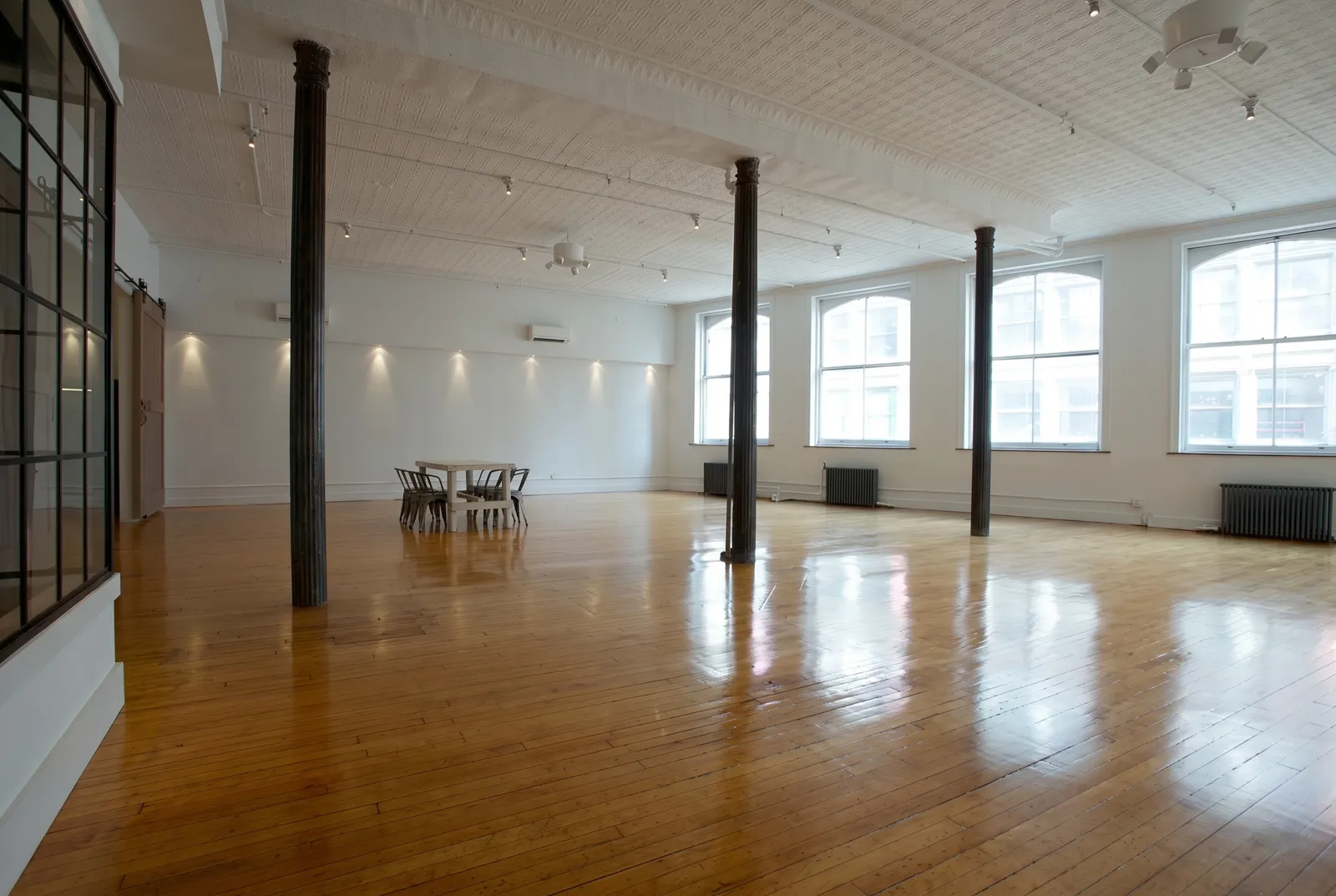 A large industrial chic room has one small table off to the side on a shiny wooden floor, while the rest of the room is empty.