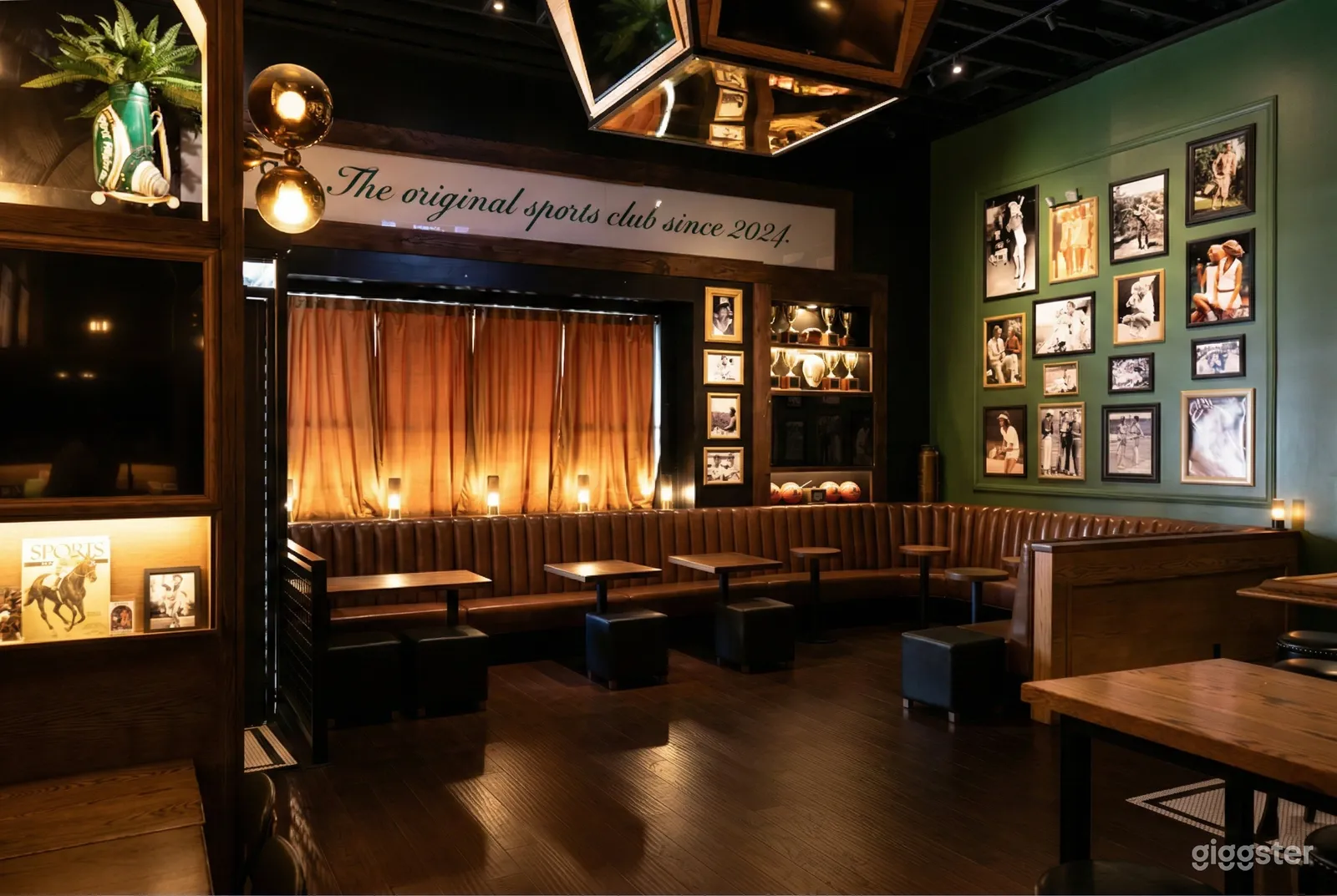 A green wall with sports photos overlooks a long curved bench and tables in a sports bar setting.