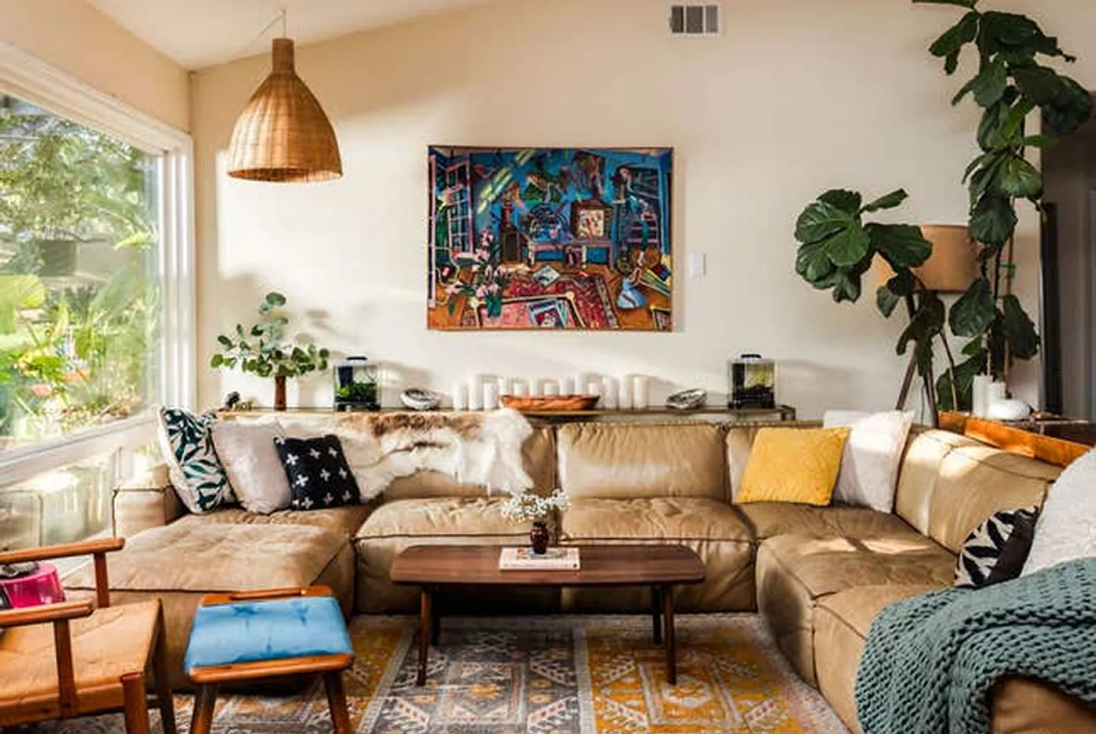 A sectional couch covered in pillows and throws is centered under a modern painting, with a large window to the side.