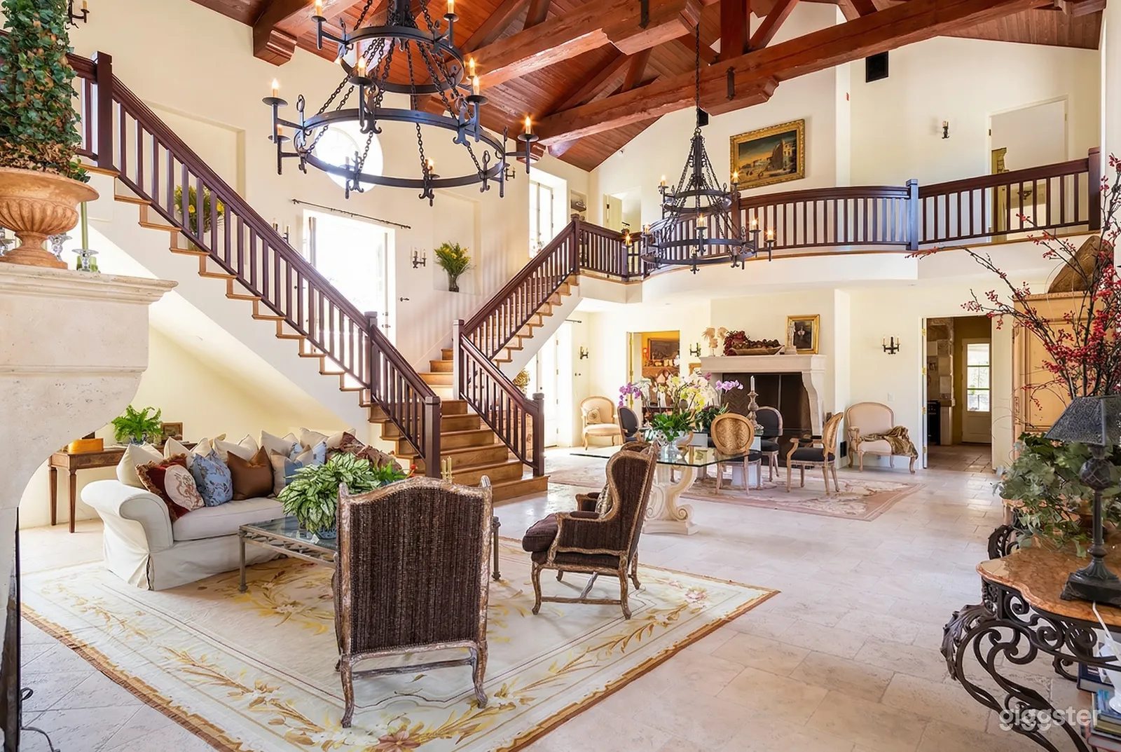 The main floor of a mansion has a dining and sitting area filled with ornate furniture and a central staircase and balcony surrounds it all.