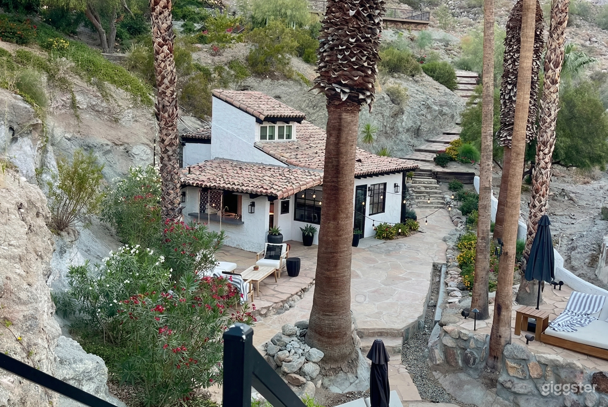 A white house with a tiled roof is nestled in a desert hill setting with desert plants and trees surrounding it.