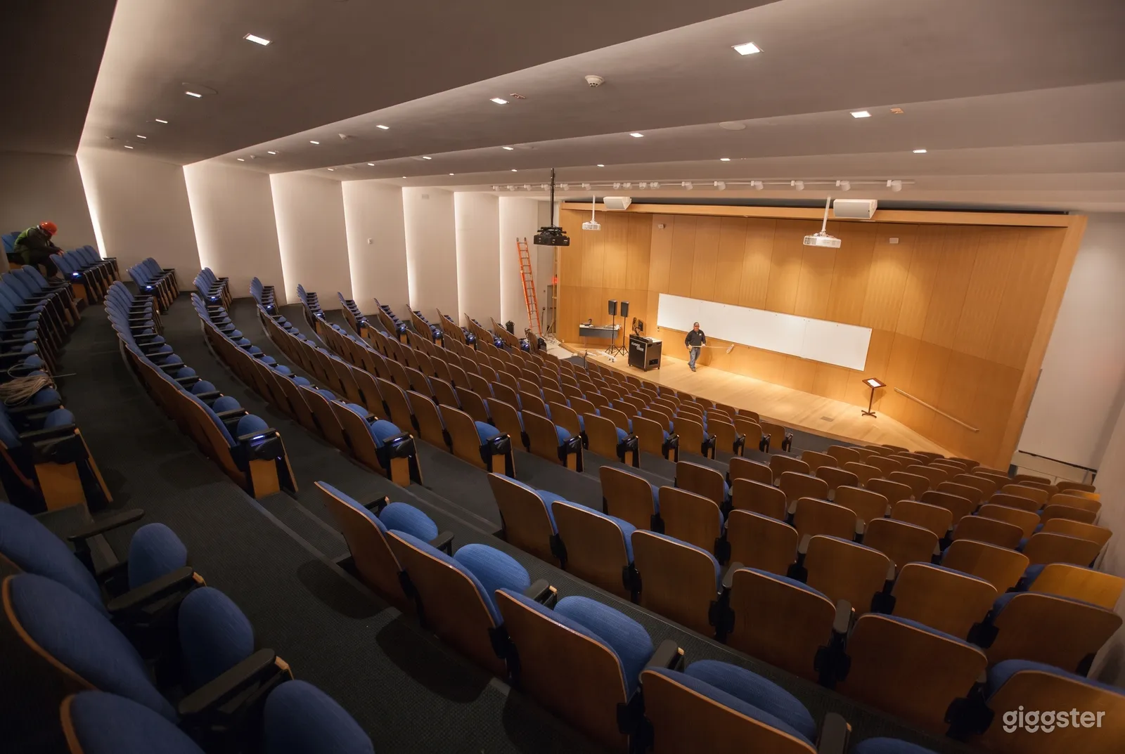 A large auditorium with rows of tiered blue seating overlooks a well-lit wooden stage area.