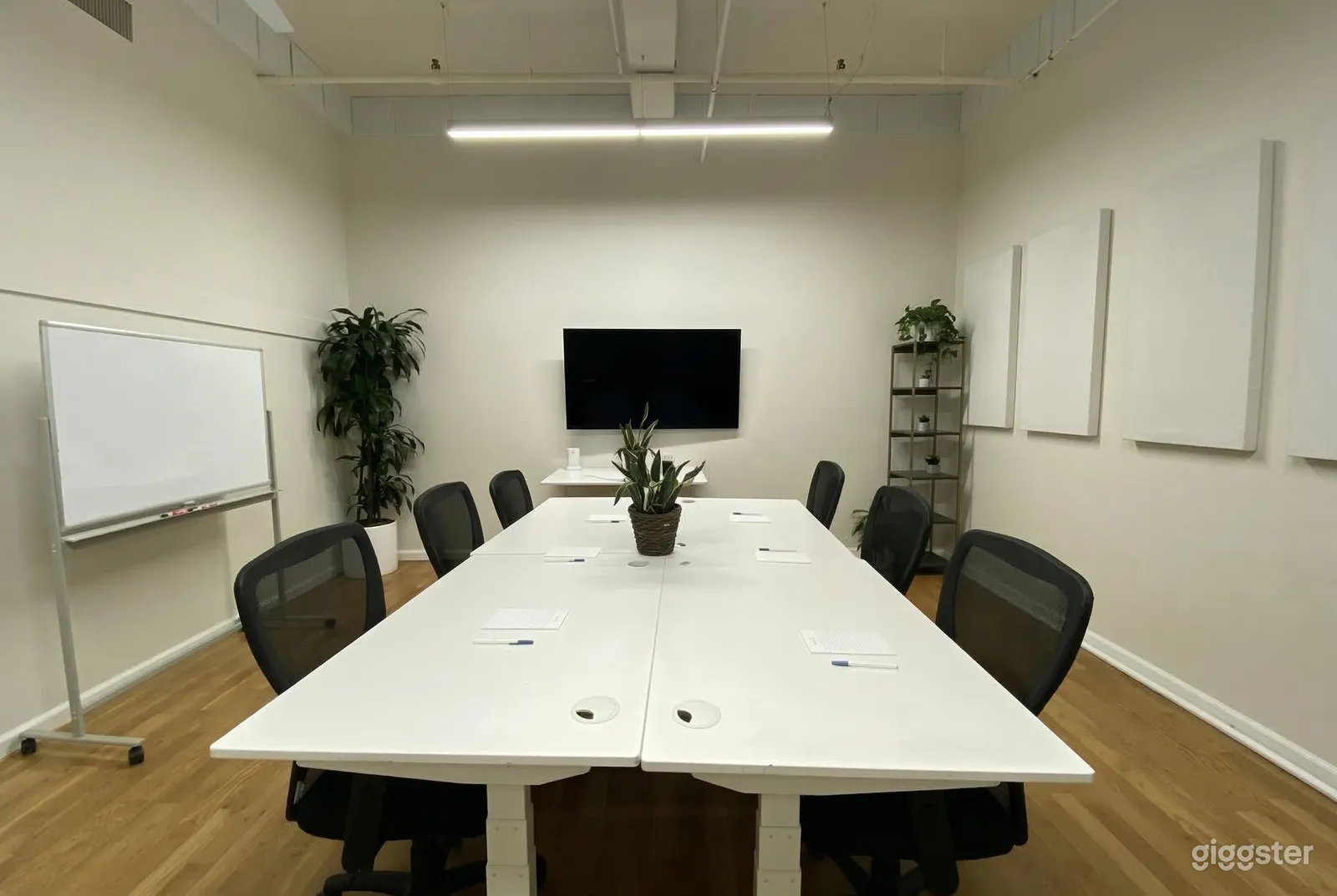 A long white conference table with black chairs is central to a space with a television and white board.
