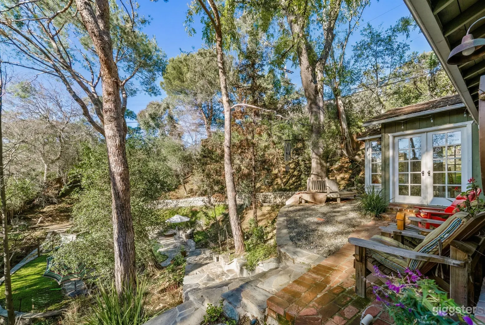 Chairs on a stone deck and walkway in front of a garden-surrounded home.