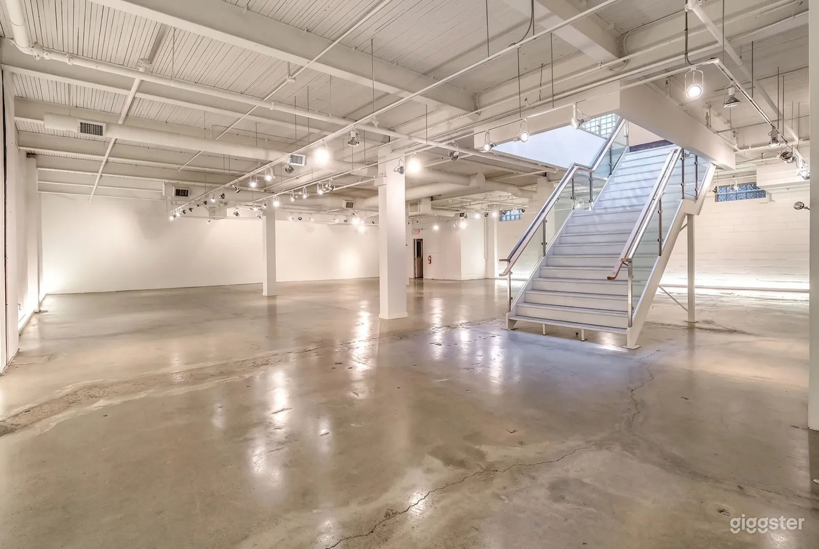 A warehouse space with glossy cement floors, white walls, a wide staircase and plenty of lighting.
