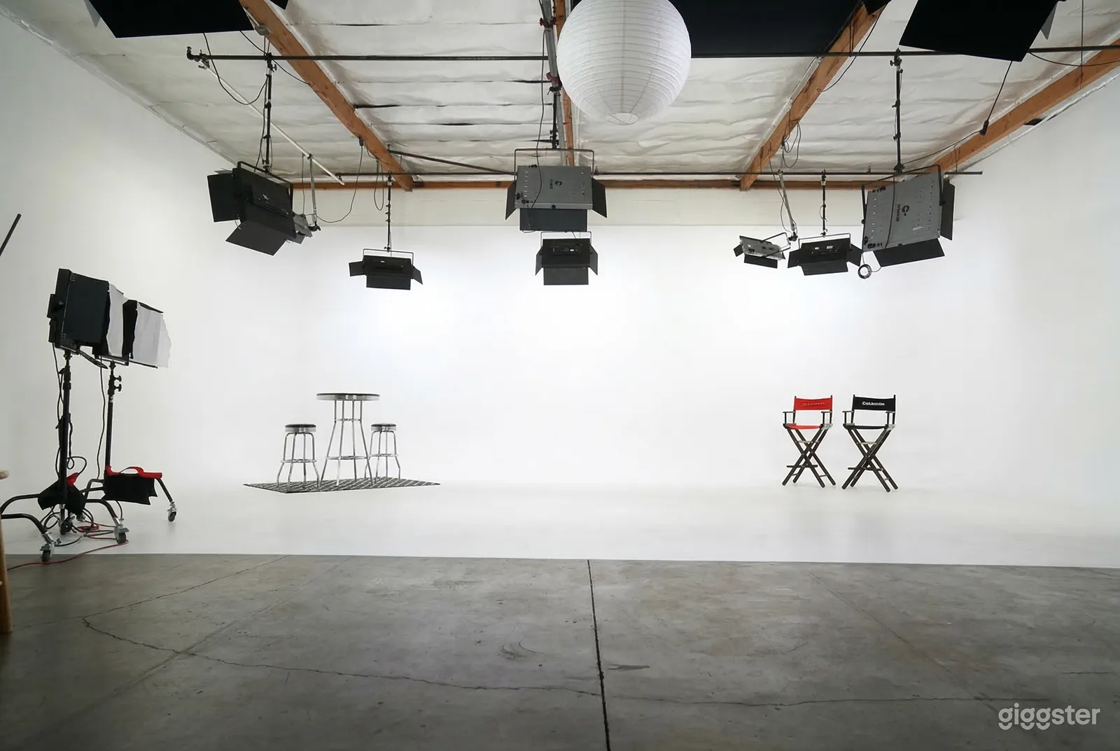 Tall chairs and benches sit in front of a floor and wall-covered white screen with supplemental lighting overhead.