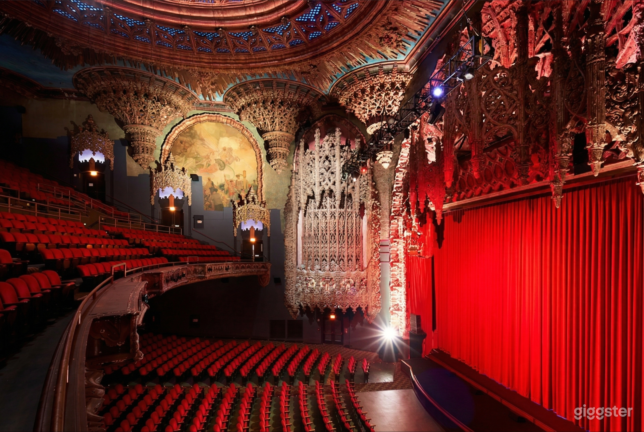 A Gothic style theater with ultra-ornate walls and ceiling has many rows of red seats facing a red curtained stage.