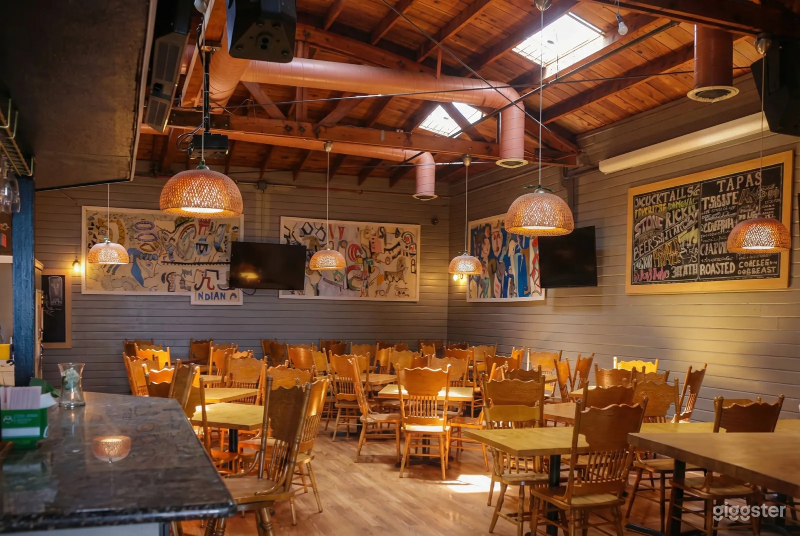 Wooden chairs and tables fill a room with an open ceiling and low hanging lights.