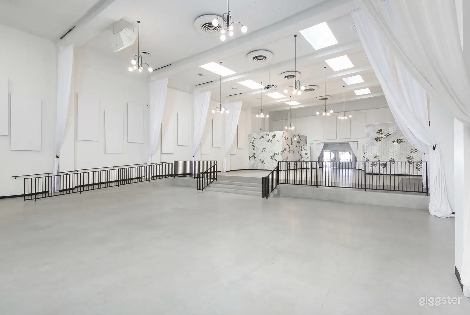 A large empty event hall is painted white, has white curtains along the walls, and low hanging white lights.