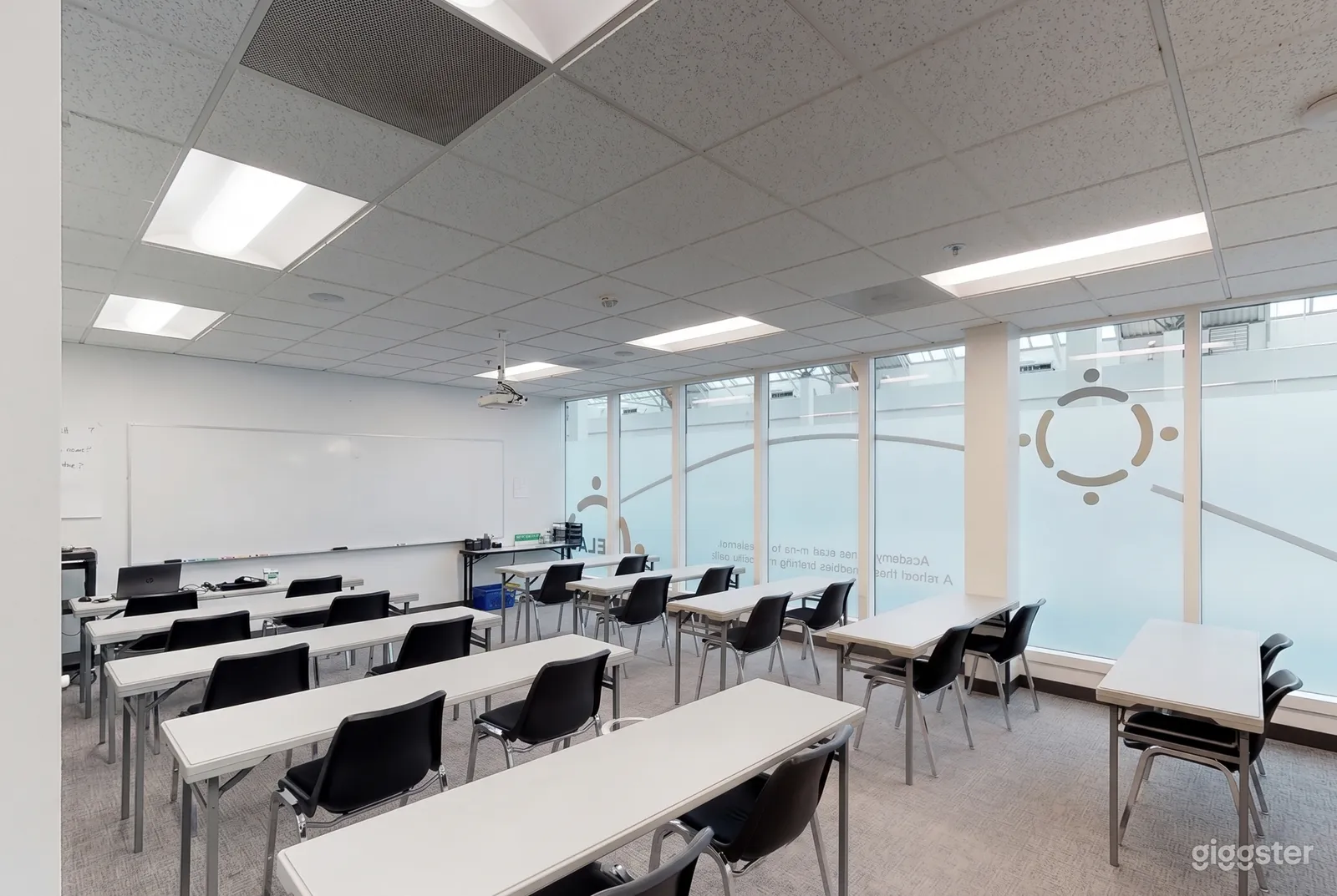 A bright lecture room has white tables and black chairs in rows, as well as frosted floor to ceiling windows.