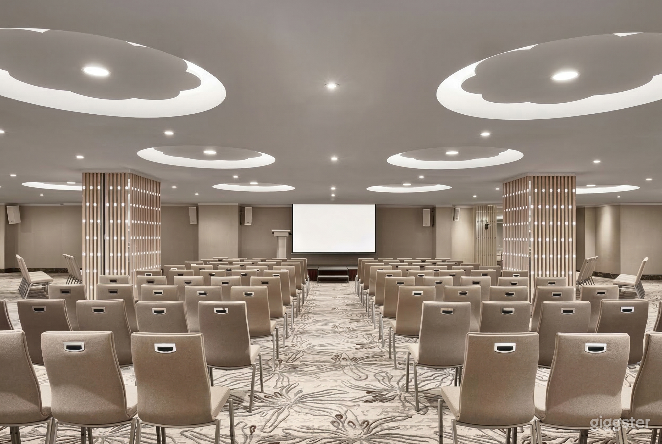 A large event room is lined with many beige chairs in rows facing a big white screen.