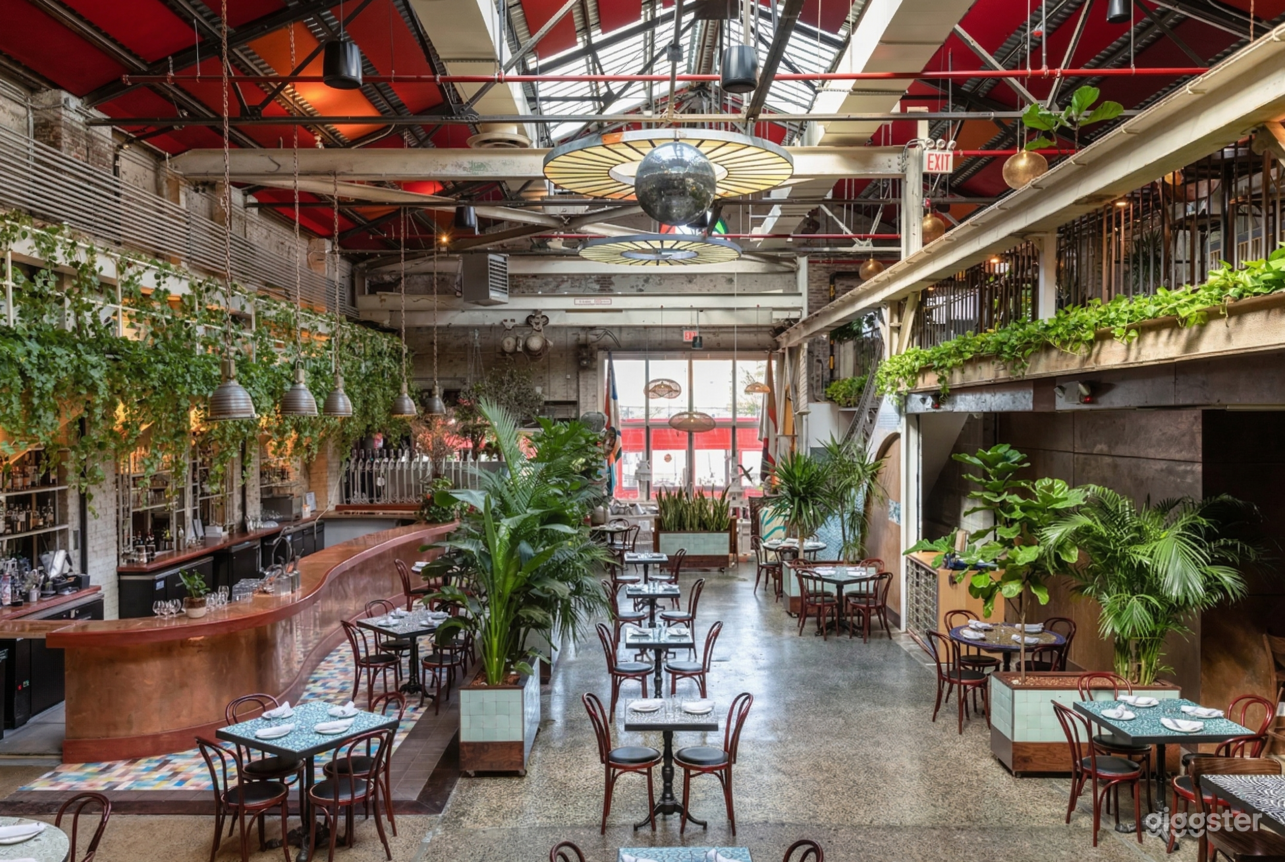 A restaurant filled with tables for two and four, with plants on the floor area and framing the walls below an industrial style ceiling.