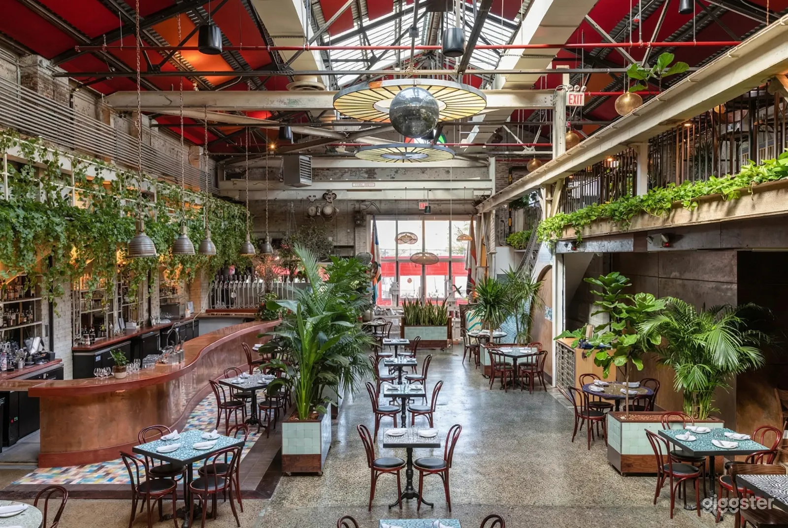 A restaurant filled with tables for two and four, with plants on the floor area and framing the walls below an industrial style ceiling.