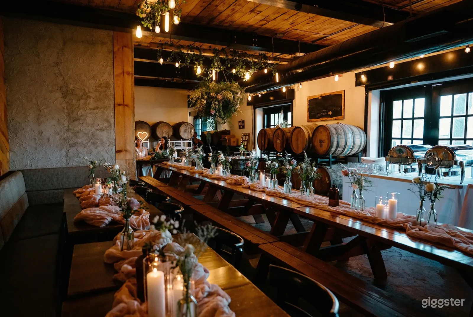 A stylish brewery room with barrels along the walls has tables decorated with runners, flowers in vintage bottles, and candles.