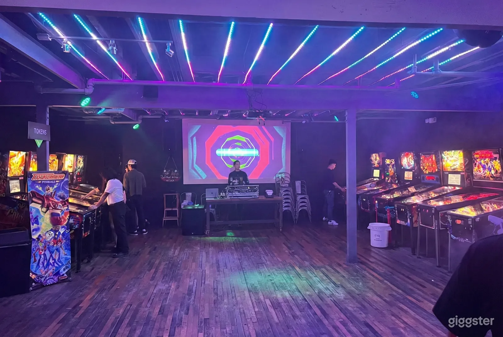 Pinball machine filled games room lit in purple with a purple and pink sign glowing in the background. 