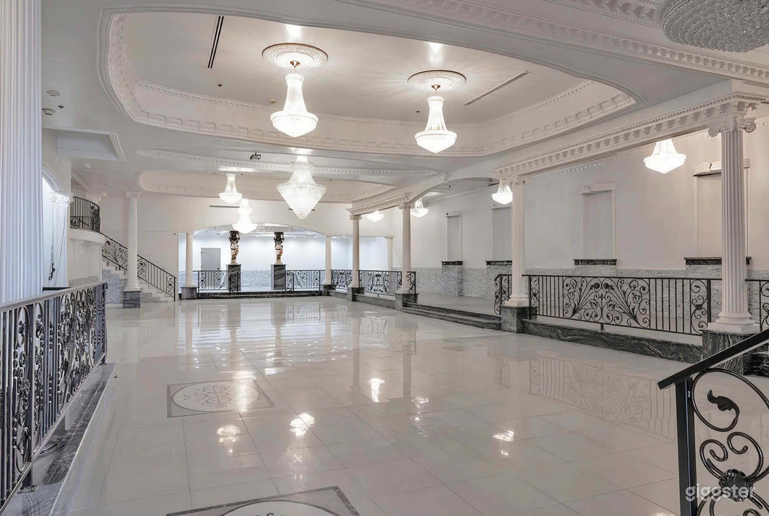 An event room with shiny ceramic floors has multiple ceiling chandeliers and is surrounded by a separate area with wrought iron fence barriers and columns.