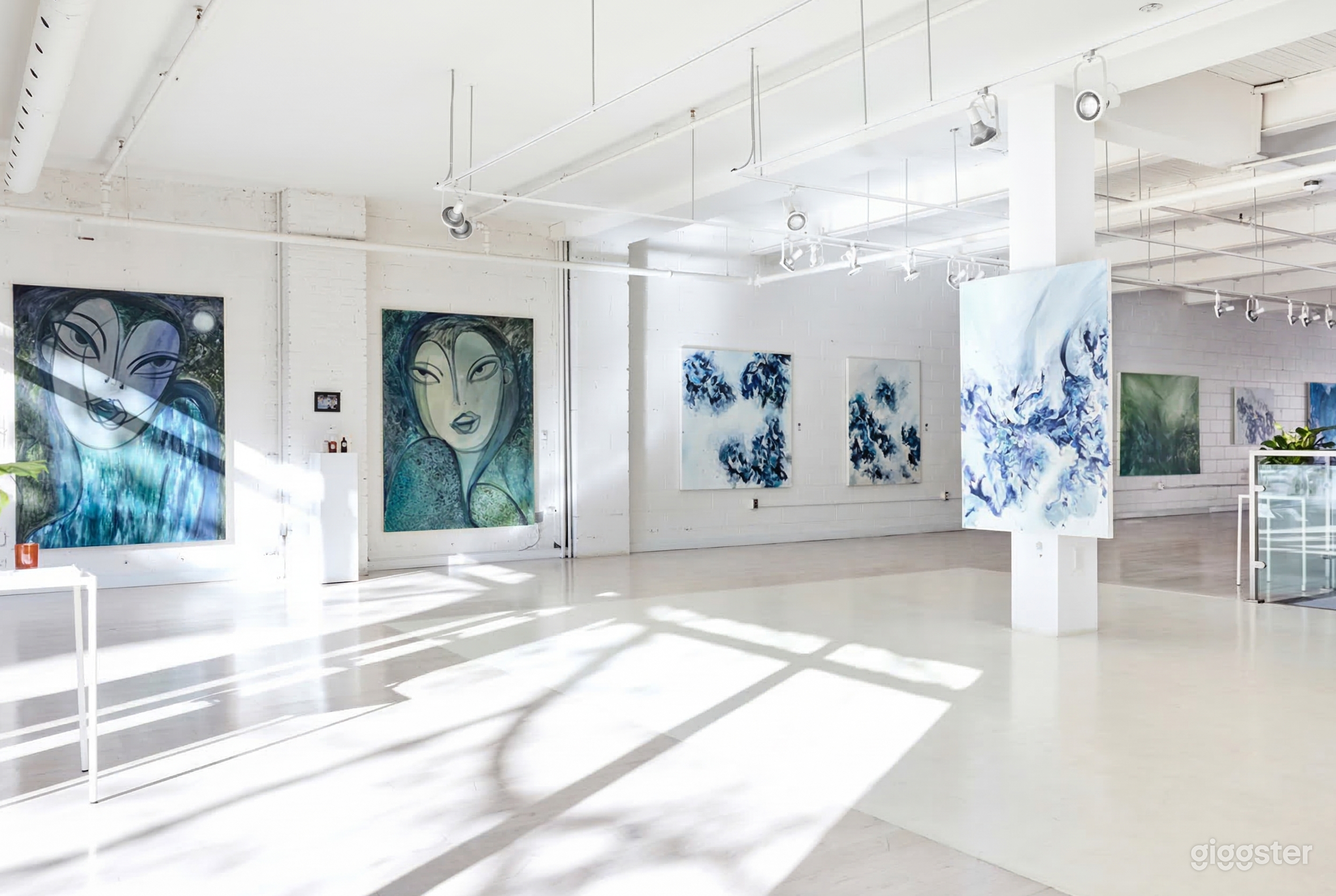 Large pieces of artwork in blue hues line the wall of a big white exposition space.