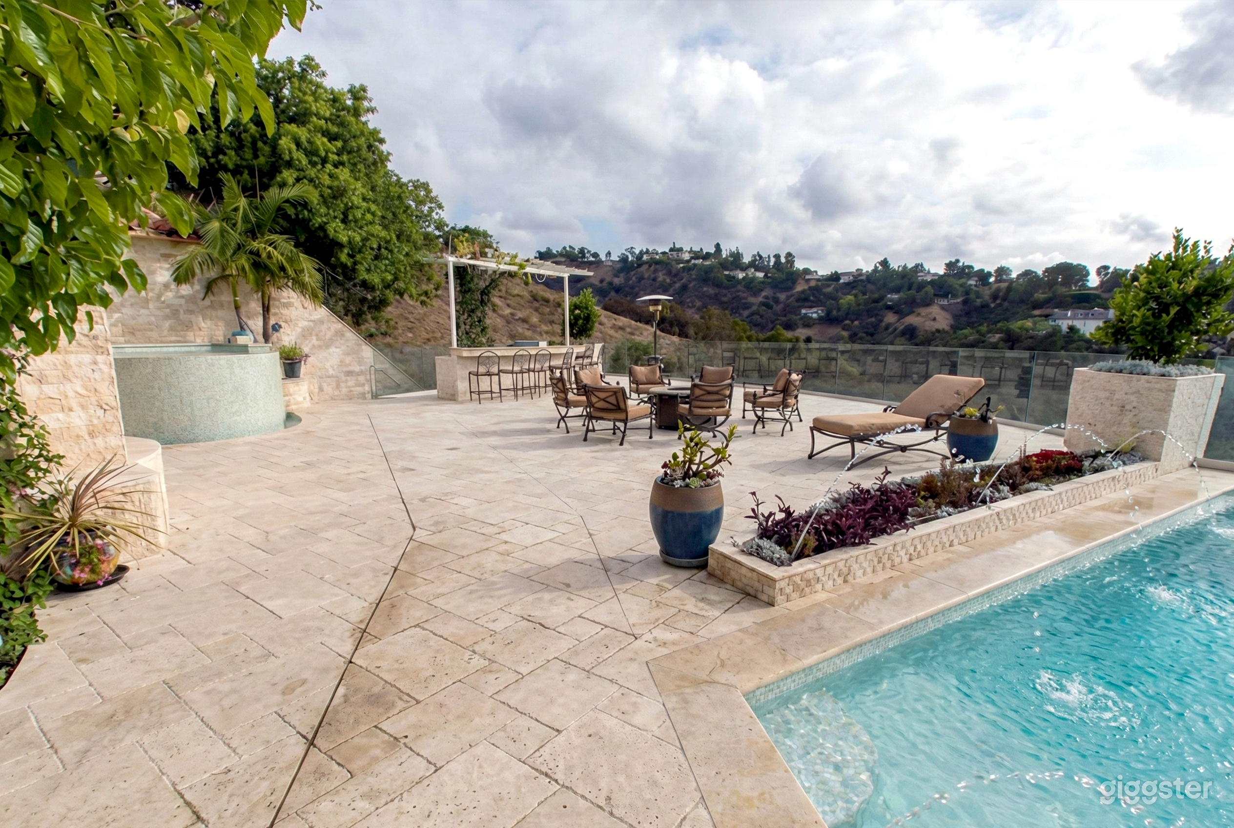 Wide open beige stone terrace and pool with fencing and hilly trees in the background.