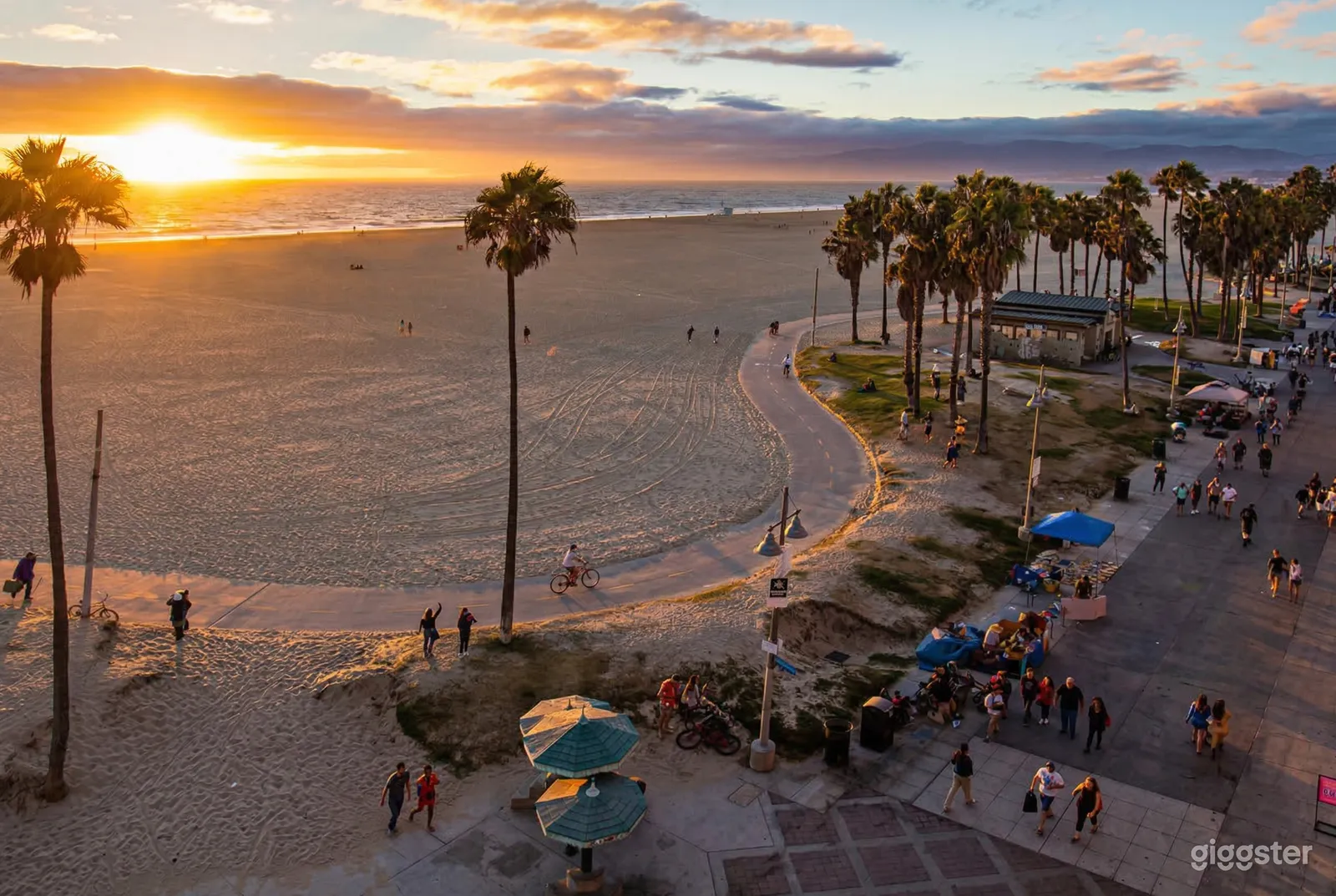An expansive sandy beach is in front of palm trees and a people-filled cement boardwalk.