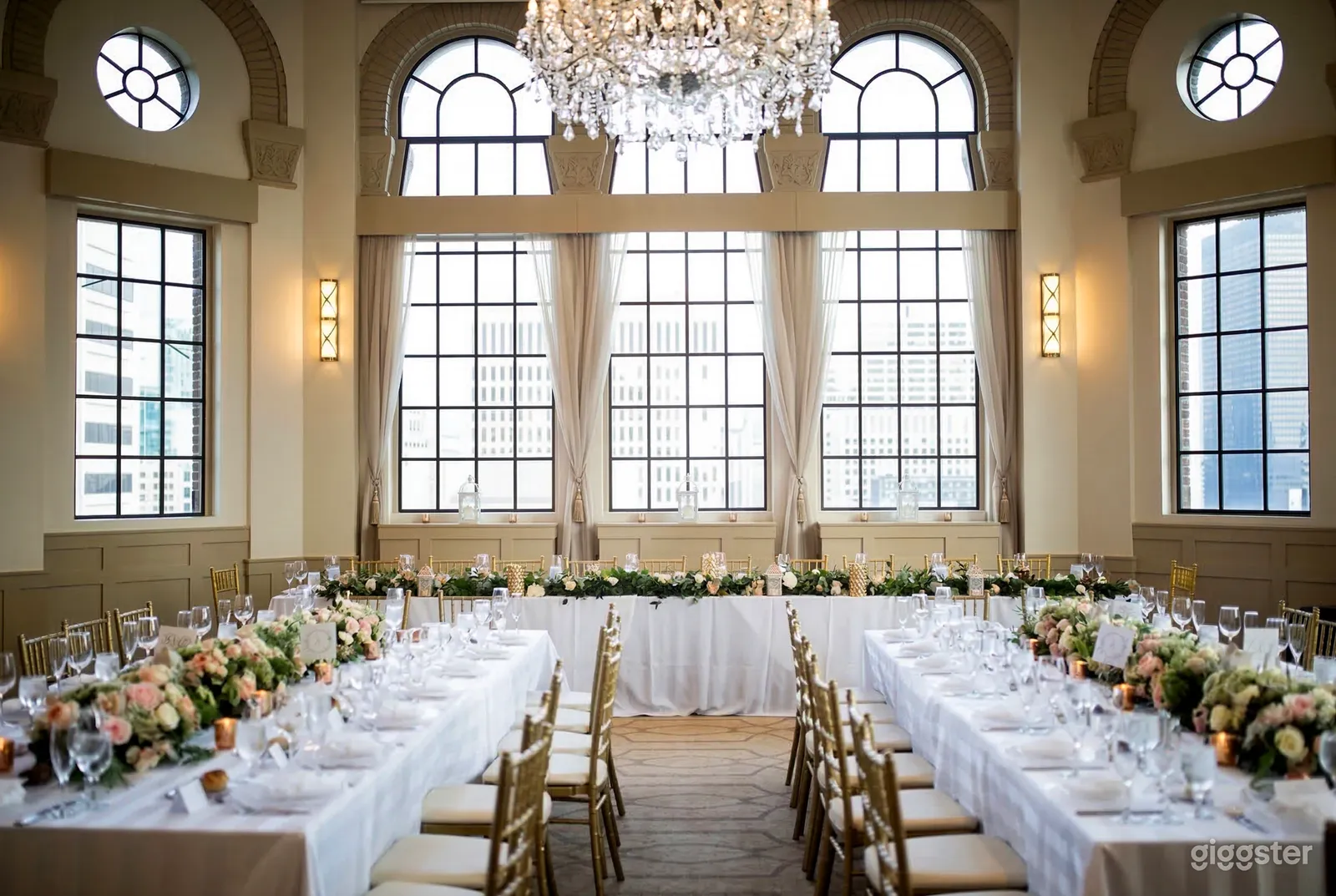 White tables in a U-shape are decorated with flowered centerpieces in front of large ornately styled windows.