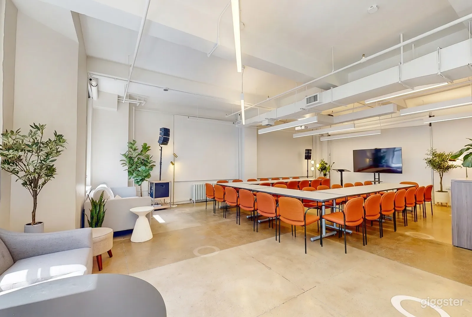 A u-shaped table with orange chairs sits in a front of a screen and podium in a modern looking classroom.