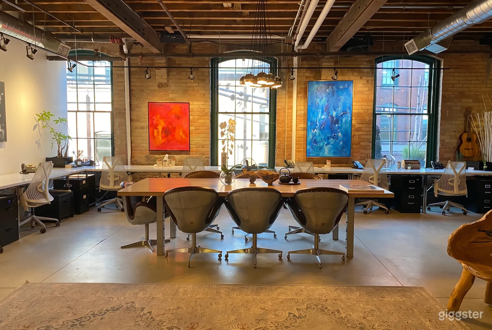 A conference table and chairs sits in front of a brick wall with colorful paintings and large paned windows.
