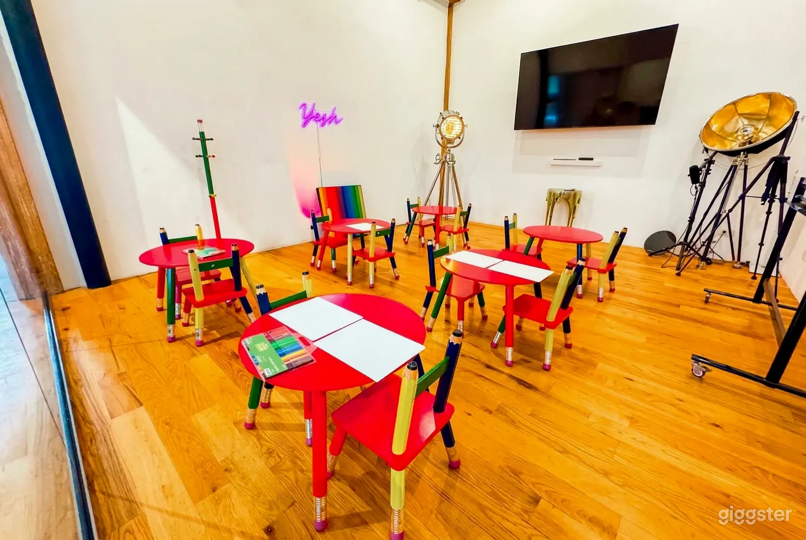 Round red tables with chairs are set up in a room with a wooden floor and large TV on the wall.