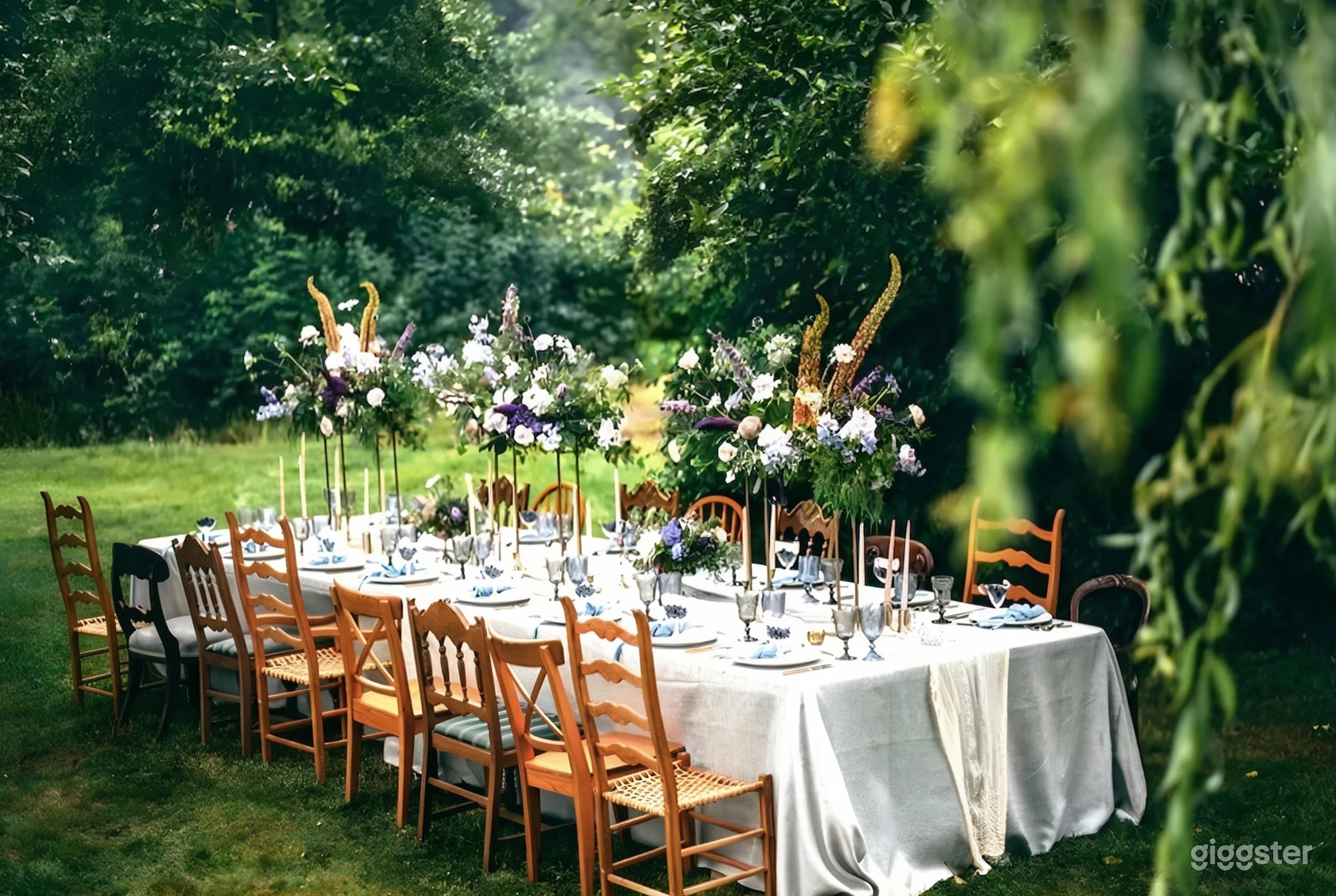 A table with various style chairs sits in a garden and is decorated with smoky colored wine glasses and large vases of wildflowers.