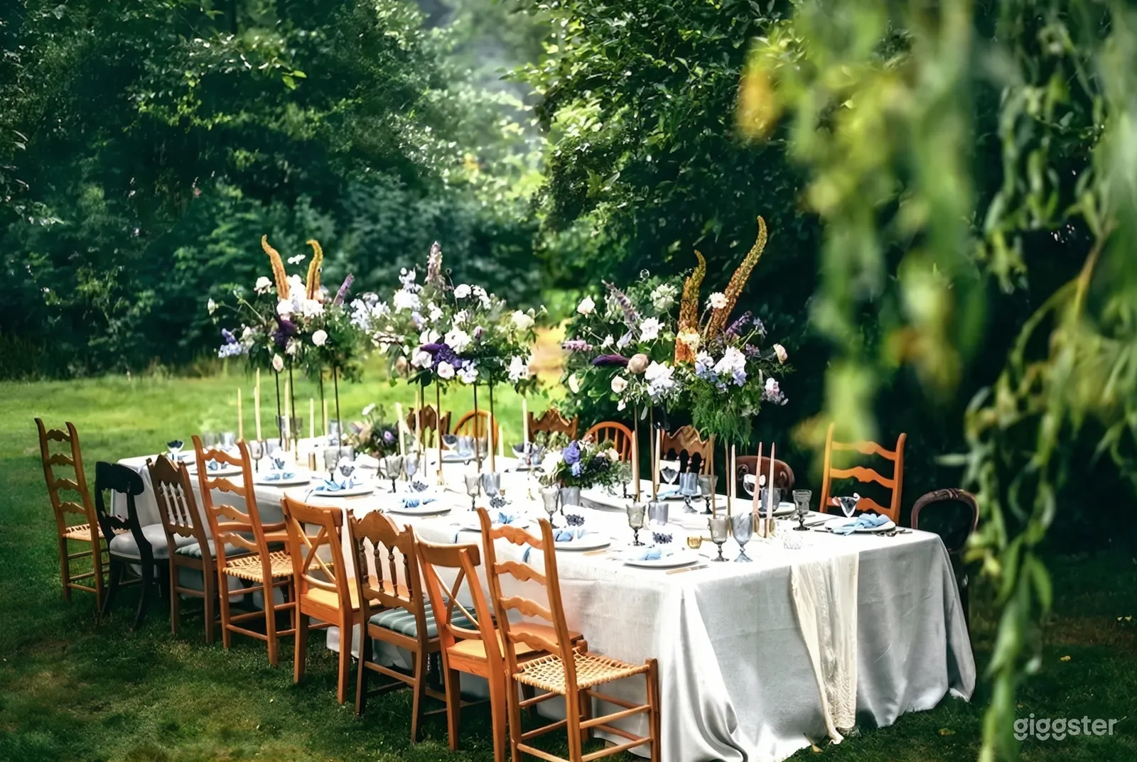 A table with various style chairs sits in a garden and is decorated with smoky colored wine glasses and large vases of wildflowers.